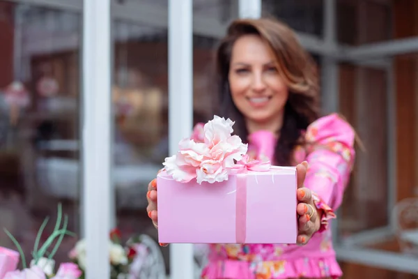 Smiling glorious brown-haired woman showing holiday present wrapped in pink box on birthday, Valentines Day or 8 march against glass doors of flower shop. Advertisement for the best gift for women