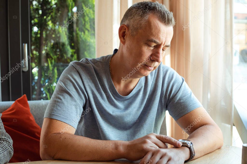 Portrait of thoughtful elderly man wearing grey T-shirt, sitting at table on sofa near window on sunny summer day at home, looking at watch, waiting for meeting. Expectation, date, business meeting.
