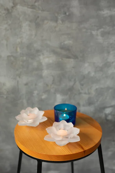 Composition of two pink candlesticks in form of beautiful flower lotus made of glass crystal and little candle burning in blue glass cup put on wooden stool with iron legs on grey marble background.
