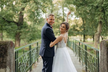 Cheerful, chancy young married couple of man and woman in glasses standing together on bridge in park, holding hands on wedding celebration. Holiday in nature, new family, newly wedded 