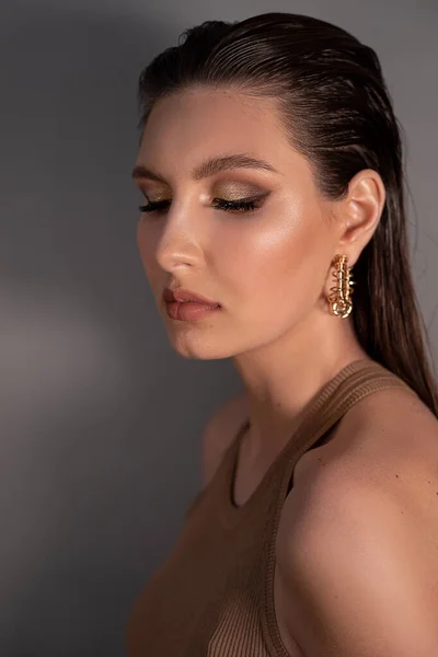 Side view of young wonderful woman with long dark hair, bright shining make-up, wearing golden earrings with closed eyes, posing on grey background. Studio shot, vertical. Beauty, cosmetics, fashion.