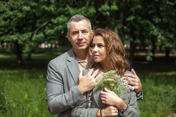Portrait of cheerful family in grey jackets, standing in park in summer. Confident elderly man father hugging embracing attractive young woman daughter holding bouquet of camomiles. Relationship.