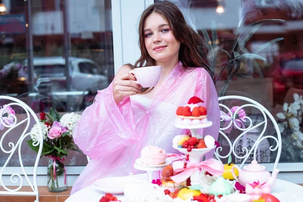 Pretty teenage girl in pink raincoat joying, tasting sweet berry dessert cakes on stand and drinking tea on the table in sidewalk outdoor cafe near glass window and flowers. Advertising cafe opening