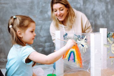 Portrait of stunning little girl sitting on art lesson, drawing white sun near rainbow with brush on stretch film wrapping around turned table legs with help of teacher in room. Creative work, crafts.