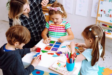 Portrait of young woman sitting at table with colored pencils, board games with pupils children playing. Teacher touching pink and white headband of little girl collecting puzzles in bright classroom.