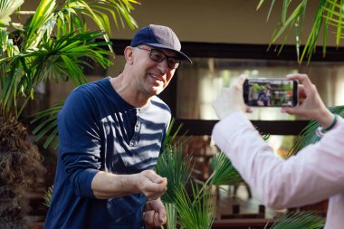Portrait of smiling middle-aged man blogger traveller stand at palm trees in cafe, gesturing, telling stories, sharing professional skills, recording educational video with smartphone held by woman.