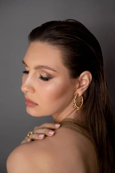 Side view of young stunning woman with long dark hair, bright golden make-up, wearing golden earrings with closed eyes, touching shoulder on grey background. Studio shot, vertical. Beauty, cosmetics.