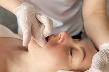Close-up of masseuse massaging face of young woman lying on couch in spa salon. Doctor thrusting finger in glove into mouth during procedure, tightening facial muscles. Anti-aging treatment, beauty.