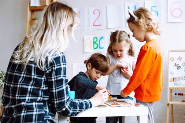 Two pretty little girls standing at table in bright classroom. Little boy sitting, playing board games. Young blond woman teacher helping children to play. Preschool education, early development.