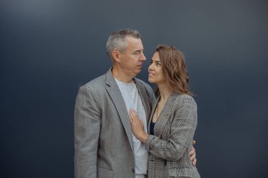 Young loving woman and handsome aged gray haired partner embracing and looking to each other, gray background. Romantic relations of beautiful couple. Happy family, true love, feelings and passion.