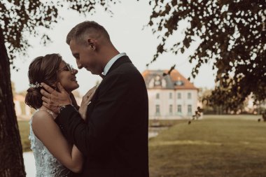 Close up portrait of smiling, glad, joyous married couple of young woman in glossy dress and glasses and groom in love embracing face to face on wedding celebration against new house. Copy space