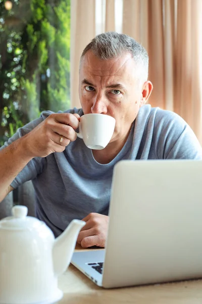 Aged attractive business man sitting in cozy cafe, drink tea and working on laptop. Implementation of modern technologies into urban lifestyle. Remote job, working time, freelance, busy people.