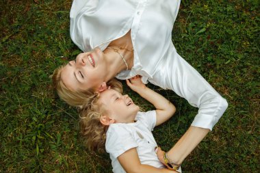 Top view of happy laughing family with closed eyes lying on grass in summer, having fun. Young attractive woman mother resting, relaxing with cute little girl daughter with curly hair. Relationship.