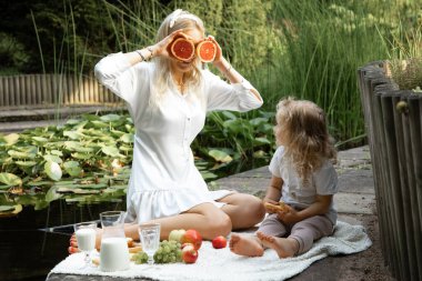Portrait of cheerful family of mother and little daughter sitting on white plaid with fruits, jug of milk on edge of pond with water lilies in summer. Young woman holding grapefruit near eyes. Picnic.