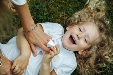 Top view of amazing laughing little girl daughter with long fluffy curly fair hair wearing white T-shirt, lying on green grass tickled by woman mother in summer, having fun, playing games, relaxing.