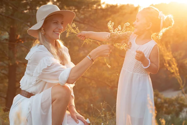 Side view of laughing family wearing white dresses, standing near pines in park forest lit up by sunset in summer. Young woman mother playing with little girl daughter using wildflowers, having fun.