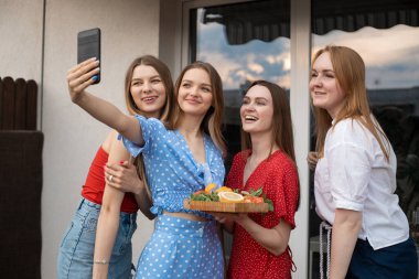 Group of young joyful wonderful women holding cut vegetables and fruits on wooden tray, stretching hand with smartphone, making selfie, enjoying moment. Friendship, celebration, hen-party, technology.