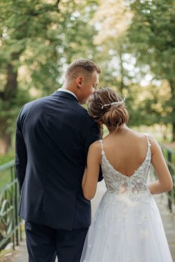 Rearview of vertical cheerful, chancy young married couple of man and woman standing together on bridge in park, holding hands on wedding celebration. Holiday in nature, new family on blurred view