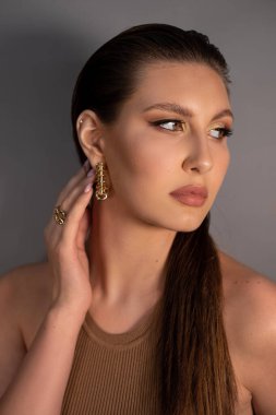 Side view of young mysterious woman with long dark hair, shining make-up, wearing golden earrings, ring, touching neck, posing on grey background. Studio shot, vertical. Beauty, cosmetics, fashion.