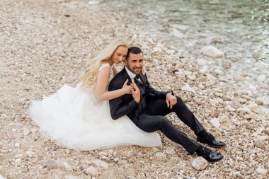 Top view of smiling wedding couple sitting on stones pebbles at shore near Adriatic sea. Young woman bride with long fair hair wearing long white wedding dress hugging man groom. Wedding, honeymoon.