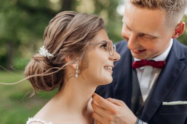 Close up joyful, laughing married couple of groom and bride in glasses on wedding celebration touching each other and stand together. Close romantic relationship, newlywed family in nature. Side view