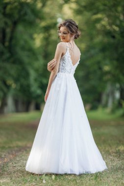 Rearview vertical portrait of young pretty, shiny bride in eyeglasses turn back in white glossy lace wedding dress posing in natural parkland outdoors. New beginnings, fashion holiday outfit concept