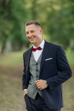 Vertical portrait of laughing, confident handsome smiling blond businessman or groom in formal tuxedo suit with red bow tie on natural blurred green background. Holiday and corporate celebration event