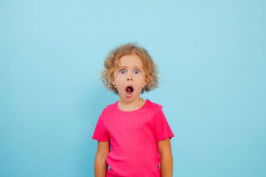 Little expressive, shocked, stressed, emotional curly blonde girl in pink shirt posing with open mouth to camera on blue isolated studio background. Advertisement for big sales for kids. Copy space