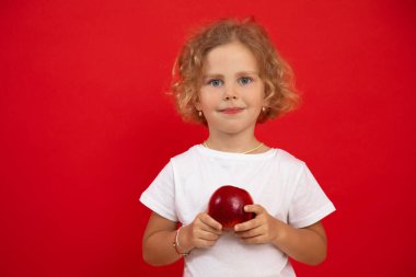 Small little expressive, positive curly blonde girl holding red juice apple in hands looking at camera on red isolated studio background. Healthy eating, lifestyle and children care. Copy space