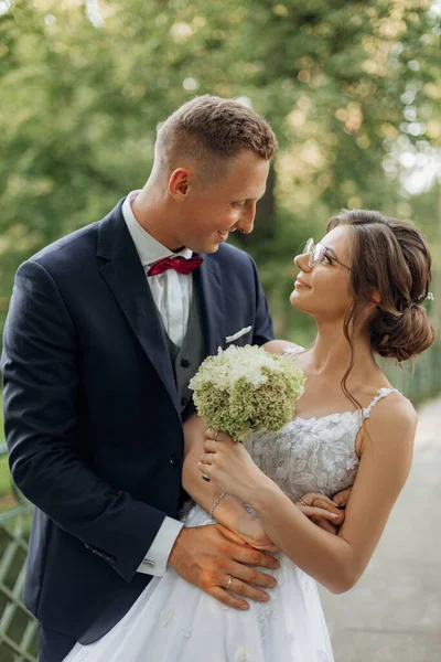 Close up vertical smiling, shy, glad newly married couple of young woman in lace dress with bouquet and groom in suit embracing and looking with love at each other on wedding celebration in nature