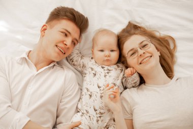 Top view of young happy smiling family in white clothes with wonderful little plump grey-eyed baby infant toddler lying on white cotton bed. Child looking seriously. Bedtime, relaxation, family, love.