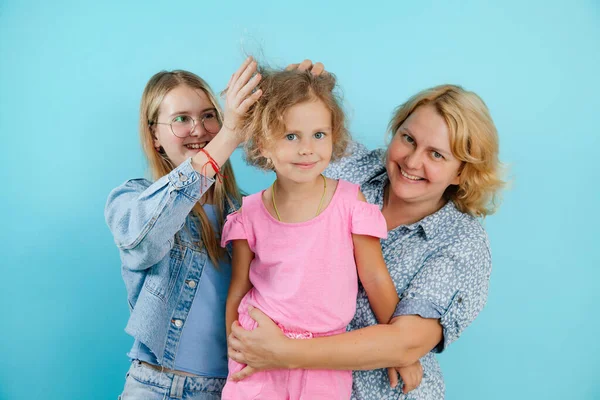 Portrait of happy family from three persons having fun on empty blue background, free copy space. Older sister ruffle hair of younger pretty sister, mom tickle belly of nice daughter.