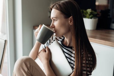 Young brown haired female drinking coffee and holding white laptop in the light kitchen home. Online exams, preparing for meeting. E-learning, new occupation and hobby, remote work and education
