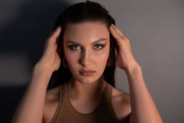 Portrait of young beautiful green-eyed woman model with bright golden make-up, pink lips titivating long dark hair, posing on grey background. Studio shot. Beauty, cosmetics, fashion, skin care.
