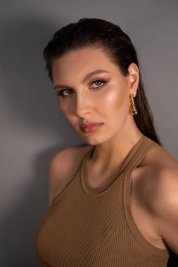 Side view of young well-groomed confident woman with long dark hair, bright shining make-up, wearing golden earrings, posing on grey background. Studio shot, vertical. Beauty, cosmetics, fashion.