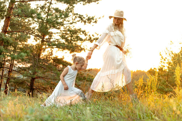 Portrait of happy family wearing white dresses, walking on tall grass among trees in park forest illumined by sunset in summer. Young woman mother holding hand of little girl daughter. Relationship.