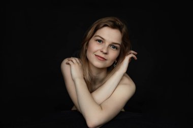 Portrait of positive, cheerful, glad and smiling young naked blonde woman look camera on black isolated background. Female covering with arms, natural beauty and face care. Waist up view. Copy space