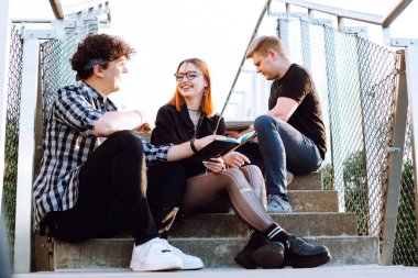 Cheerful talking relaxing and joyful small group of people, two men and red hair woman in glasses sitting on stairs and reading book. Three young friends on meeting weekend, vacation. Hipster culture
