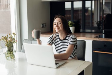 Laughing and joyful dark haired businesswoman sitting in the kitchen, working on white laptop and drinking coffee near big window. Wireless technology, video call, internet on remote job, E-commerce