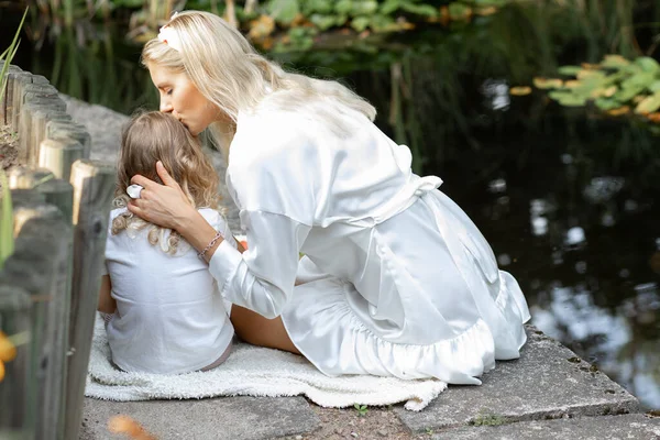 Back view of wonderful family sitting on white plaid on edge of pond with green water lilies near wooden fence in summer. Young woman mother kissing little girl daughter. Picnic, relationship, love.