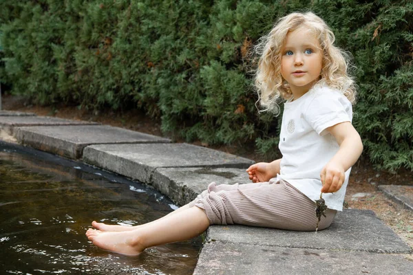 Portrait of cherubic blue-eyed little girl with long curly fluffy hair sitting on concrete edge of pond swimming pool with bare feet in water near bushes, playing in garden park, holding dry frog.