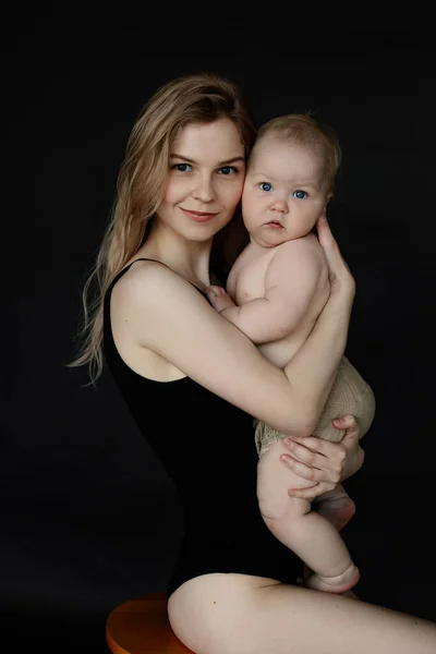 Vertical portrait of young blonde glad mother hugging little baby in arms, look at camera on black background. Children love and care, spending time together. Offspring development. Maternity vacation