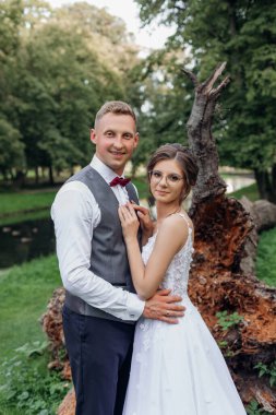 Vertical portrait of fortunate, blissful married couple of husband hugging wife after wedding celebration, looking at camera and standing near big tree stem in green natural parkland outdoors. 