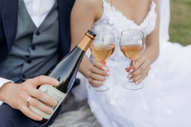 Close up cropped unrecognizable groom and bride hands drinking and pouring bubbly champagne wine from bottle and glasses together, sitting on blanket in park and having fun on wedding celebration day.