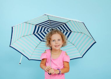 Small little expressive, smiling curly blonde girl in pink shirt hold striped umbrella in hands, look up on blue isolated studio background. Safety and children care, protection, insurance. Copy space