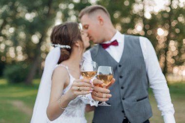 Happy married couple of bride in wedding dress and groom in suit kissing and drinking bubbly champagne together, cheers and having fun on wedding celebration day in natural parkland outdoors.