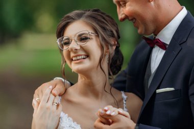 Charming, laughing, romantic stylish young married couple of man in suit and woman in glasses and white wedding dress embracing each other in nature. Happy marriage, traditional family in park
