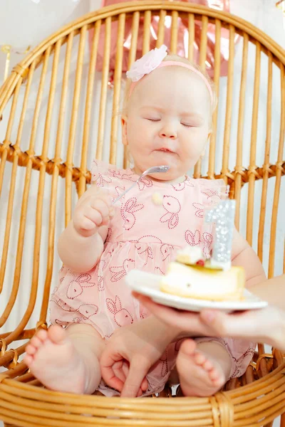 ピンクのドレスと頭の上の花で目が近い垂直裸足の幼児の子供は ウェブベッドチェアに座って誕生日ケーキを試飲します 1年だ カップケーキとキャンドル付きの作物の手を保持プレート — ストック写真