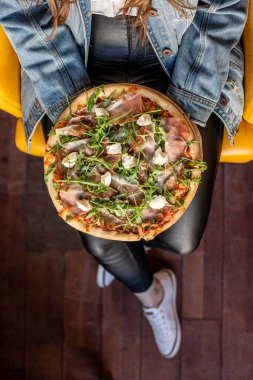 Dikey doğranmış, tanınmayan bir kadın elinde yeşil peynirli, pastırmalı lezzetli mozzarella pizzası, kafede sarı sandalyede oturan et. Tavsiye ve restoran reklamı. Üst görünüm