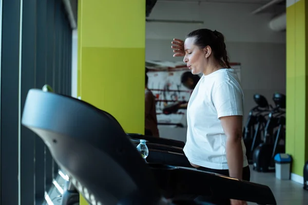 Fat woman run on treadmill at fitness gym wiping sweat from face by hand and exhale deeply through mouth, side view. Adult lady do cardio workout on exercise machine. Weight loss, sport lifestyle.